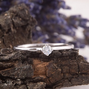 Pu&ograve; includere: Un delicato anello d'argento con una singola gemma rotonda e trasparente. L'anello &egrave; posto su un pezzo di legno scuro e strutturato, con uno sfondo sfocato di fiori viola. La fascia dell'anello &egrave; sottile e lucida.