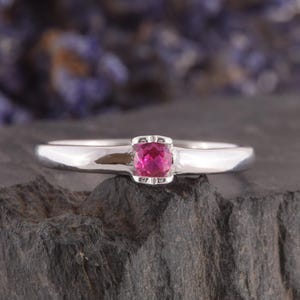 Può includere: Un anello in argento lucido con una singola pietra preziosa rotonda di colore rosa vivace, incastonata in una semplice montatura a quattro griffe. La fascia è liscia e metallica, con un design pulito che esalta il colore della pietra.