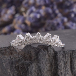 May include: A silver-toned ring featuring a curved, crown-like design adorned with five prominent round-cut clear stones along the top edge, complemented by a delicate row of smaller pav&eacute;-set clear stones running along the entire band length.