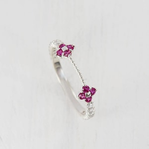 以下が含まれることがあります： 2つのピンクの花の形をした宝石のクラスターが付いたシルバーリング。リングは、2つのクラスターを繋ぐ繊細なチェーンが付いています。