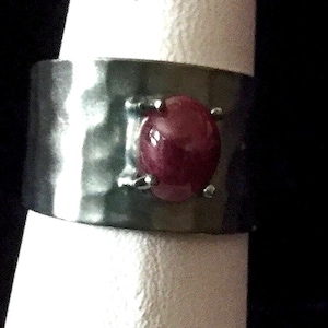 May include: A wide band ring crafted from dark, hammered metal featuring a polished, oval-shaped deep red gemstone set securely in the centre with four metal prongs. The surface of the band displays a textured, dimpled finish throughout its width.