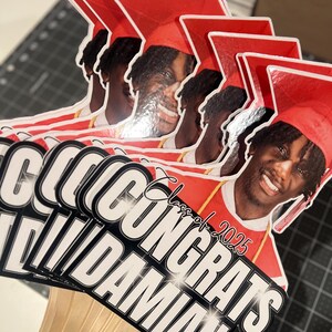 May include: A stack of graduation photo props featuring a smiling person wearing a red graduation cap and gown. The props have the text "CONGRATS DAMIAN" and "Class of 2025" in white and black.