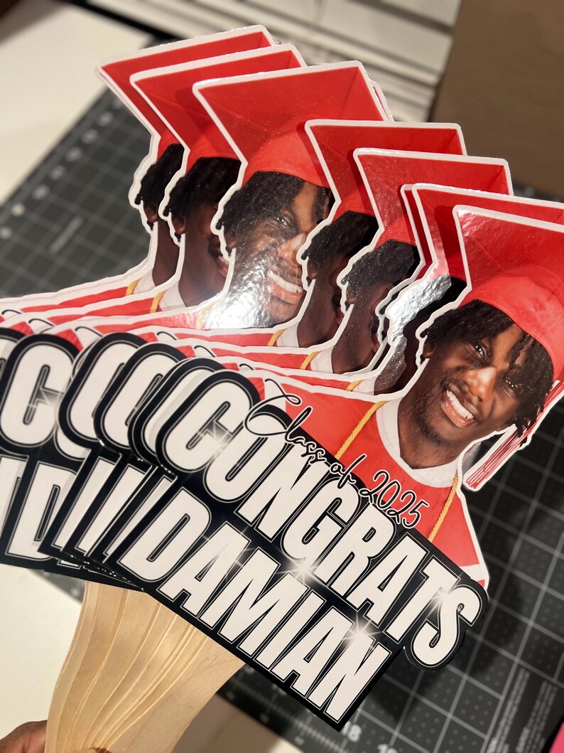 May include: A stack of graduation photo props featuring a smiling person wearing a red graduation cap and gown. The props have the text "CONGRATS DAMIAN" and "Class of 2025" in white and black.
