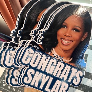 May include: A stack of graduation photo props featuring a smiling person with dark hair and a pearl necklace. The props have the text "Congrats Skylar" and "Class of 2025" in white and navy blue.