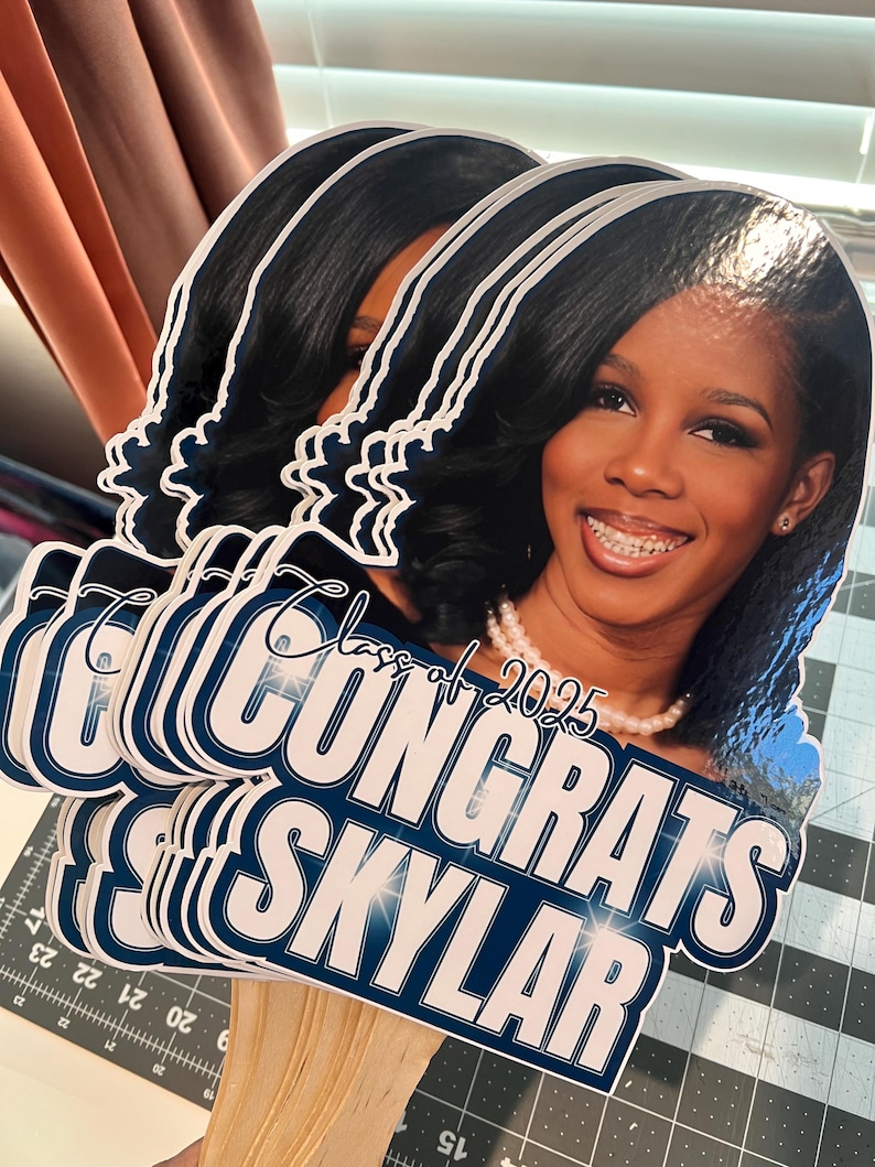 May include: A stack of graduation photo props featuring a smiling person with dark hair and a pearl necklace. The props have the text "Congrats Skylar" and "Class of 2025" in white and navy blue.