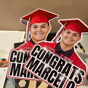 May include: Two graduation yard signs with the text "Congrats Marcelo" and "Class of 2024". The signs feature a smiling person wearing a red graduation cap and gown. The signs are held by wooden sticks.