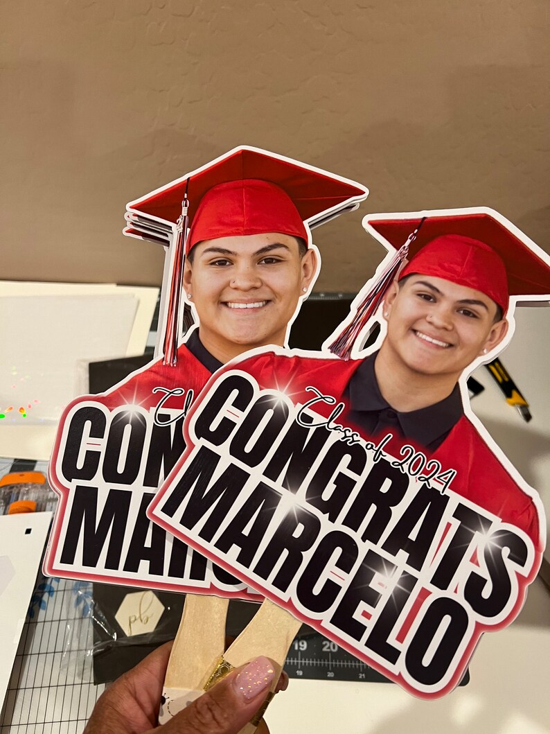 May include: Two graduation yard signs with the text "Congrats Marcelo" and "Class of 2024". The signs feature a smiling person wearing a red graduation cap and gown. The signs are held by wooden sticks.