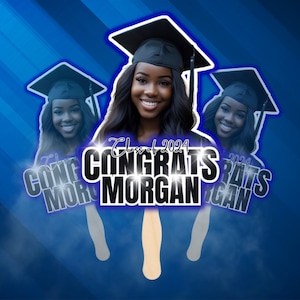 May include: A graduation fan featuring a smiling woman wearing a graduation cap. The fan has the text "Congrats Morgan" and "Class of 2024" in white and black. The background is blue with diagonal stripes.