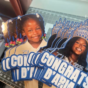 May include: Graduation photo booth props featuring smiling young women. The props have blue and white text that reads "Congrats D'Aria" and "Class of 2025". The props are held by wooden sticks.