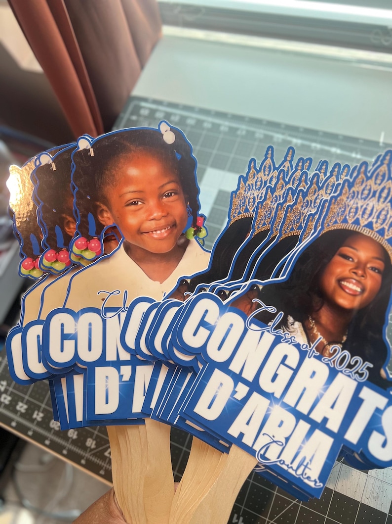 May include: Graduation photo booth props featuring smiling young women. The props have blue and white text that reads "Congrats D'Aria" and "Class of 2025". The props are held by wooden sticks.