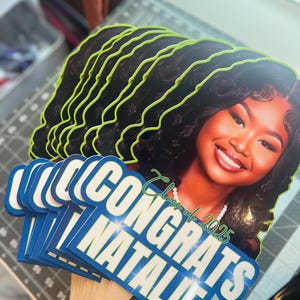 May include: A stack of graduation photo props featuring a smiling woman with dark hair. The props have a neon green outline and the words "CONGRATS NATALIE" in white on a blue background. The text "Class of 2025" is also visible.