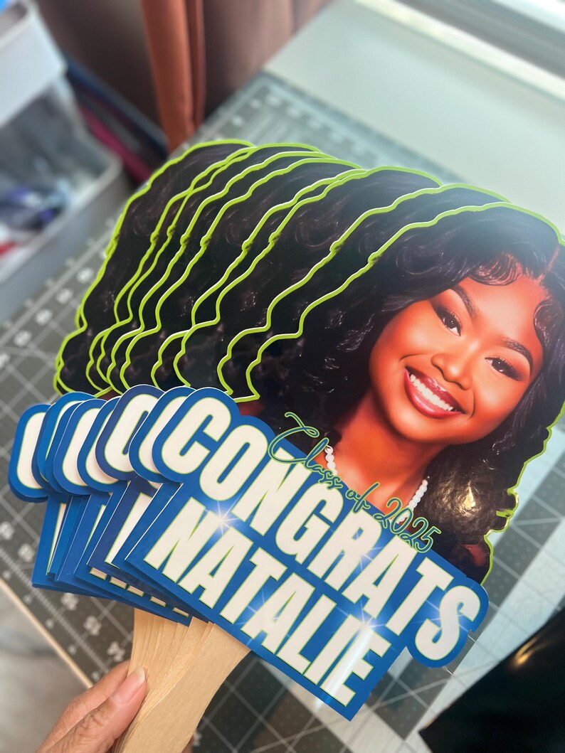 May include: A stack of graduation photo props featuring a smiling woman with dark hair. The props have a neon green outline and the words "CONGRATS NATALIE" in white on a blue background. The text "Class of 2025" is also visible.