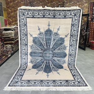 Puede incluir: Una alfombra rectangular grande con un campo de color crema y un diseño floral azul central. El borde y los elementos del diseño son en tonos azules. La alfombra tiene flecos en los extremos cortos y se exhibe en una tienda.