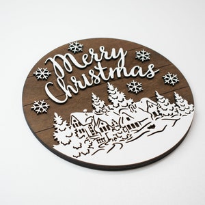 May include: A round wooden sign with a brown woodgrain background and white lettering. The sign says "Merry Christmas" and features a winter scene with houses, trees, and snowflakes.