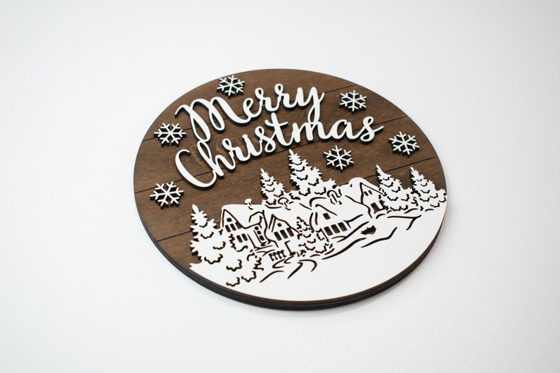 May include: A round wooden sign with a brown woodgrain background and white lettering. The sign says "Merry Christmas" and features a winter scene with houses, trees, and snowflakes.