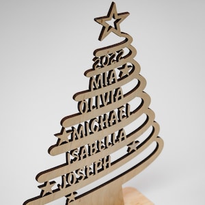 Op de afbeelding: Houten kerstboomversiering met een ster bovenop en de namen Mia, Olivia, Michael, Isabella en Joseph. Het jaar 2022 wordt ook weergegeven. De boom is gemaakt van lichtgekleurd hout met een donkerdere omtrek.