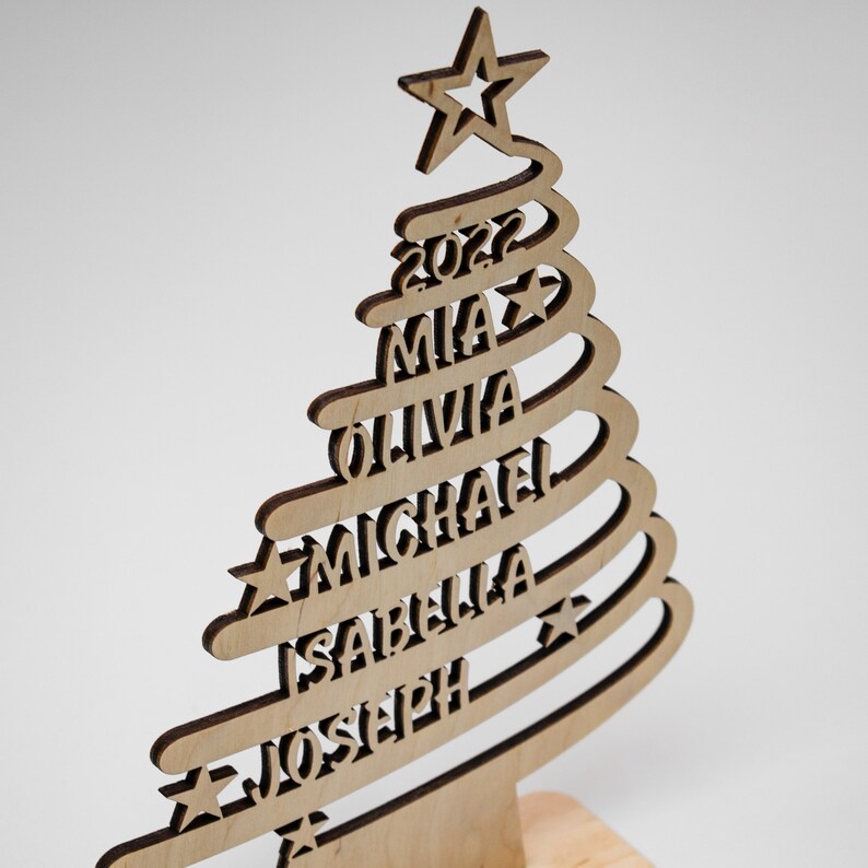Op de afbeelding: Houten kerstboomversiering met een ster bovenop en de namen Mia, Olivia, Michael, Isabella en Joseph. Het jaar 2022 wordt ook weergegeven. De boom is gemaakt van lichtgekleurd hout met een donkerdere omtrek.