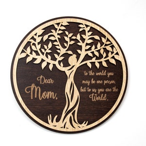 May include: A round wooden plaque with a laser-cut tree design. The tree is made of light wood and the plaque is dark brown. The text "Dear Mom, to the world you may be one person, but to us you are the World." is engraved on the plaque.