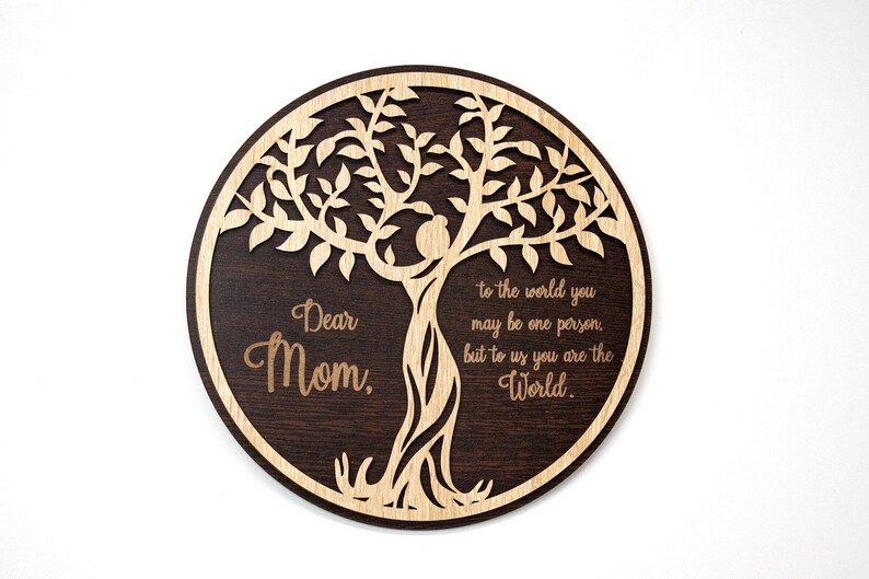 May include: A round wooden plaque with a laser-cut tree design. The tree is made of light wood and the plaque is dark brown. The text "Dear Mom, to the world you may be one person, but to us you are the World." is engraved on the plaque.