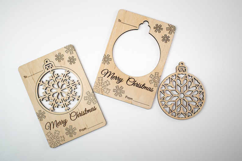 May include: Three wooden Christmas ornament gift tags with a snowflake design. The tags have a cut-out shape of an ornament and the words "Merry Christmas" and "To" and "From".