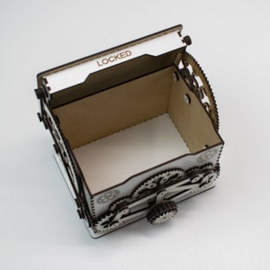 May include: A white and brown wooden box with a hinged lid, featuring a gear mechanism design. The word "LOCKED" is visible on the lid. The box has an open interior and is designed with intricate laser-cut details.
