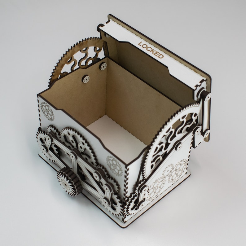 May include: A white wooden box with a hinged lid, featuring intricate gear-shaped designs and the word "LOCKED" on the lid. The box has a steampunk aesthetic, with detailed cutouts and a mechanical appearance. The box is empty.