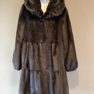 May include: A dark brown faux fur coat featuring a large, wide collar, long sleeves with gathered cuffs, and a tiered design with horizontal seams across the midsection and lower portion, creating a voluminous, textured appearance throughout.