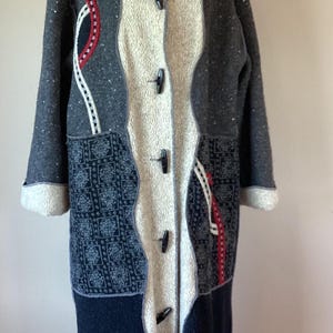 May include: A long, textured coat featuring a central cream-colored panel with four black toggle closures. The coat incorporates dark gray speckled fabric, black panels with a white geometric pattern, and red and white decorative trim accents.