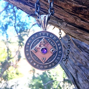 May include: A silver necklace with a round pendant featuring the words "Gratitude, Love, Joy, Faith, Peace, Compassion, Serve, Evolve" around a square with a purple gemstone in the center.