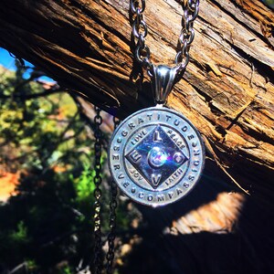 May include: Silver necklace with a round pendant featuring the words "Gratitude, Peace, Joy, Faith, Love, Serve, Compass, Passion" around the edge. The pendant has a diamond-shaped design in the center with a small crystal in the middle.