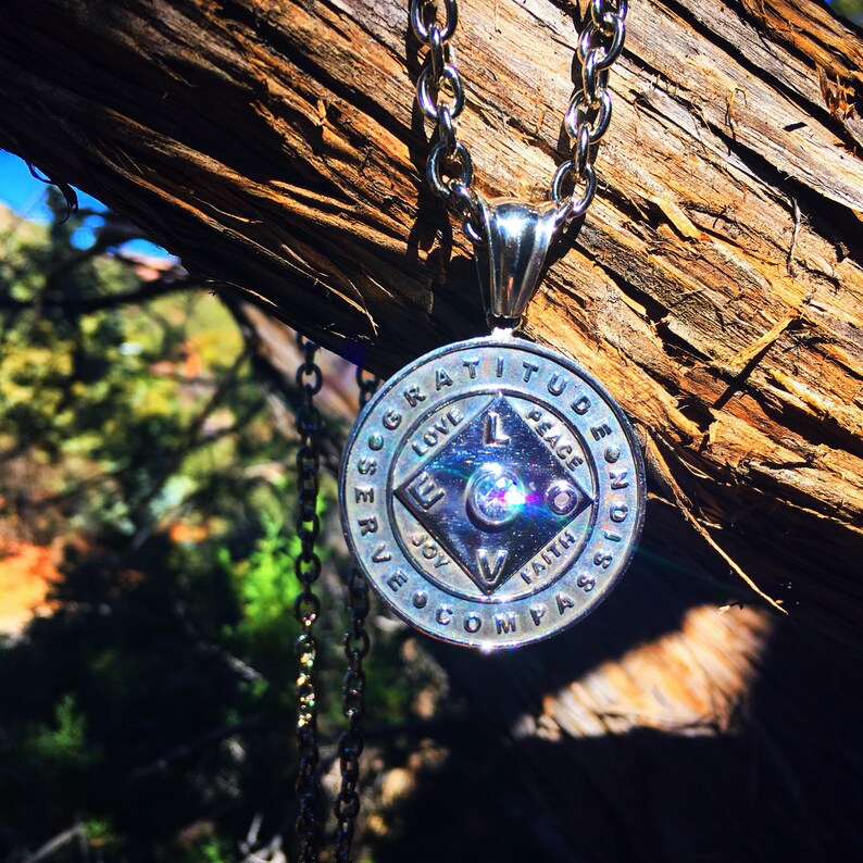 May include: Silver necklace with a round pendant featuring the words "Gratitude, Peace, Joy, Faith, Love, Serve, Compass, Passion" around the edge. The pendant has a diamond-shaped design in the center with a small crystal in the middle.
