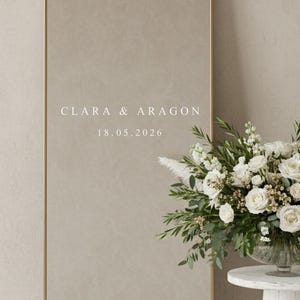 Könnte beinhalten: Ein hoher, goldener Bogen-Spiegel mit den Namen "CLARA & ARAGON" und dem Datum "18.05.2026" auf dem Glas. Ein weißes Blumenarrangement steht auf einem kleinen, weißen, runden Tisch. Zusätzliche Blumen liegen auf dem Boden.
