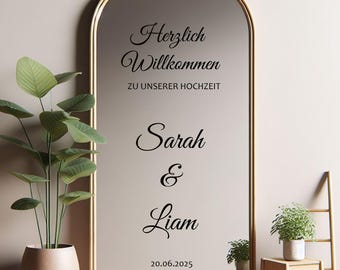 Pode incluir: Um espelho grande e arqueado com moldura dourada. O espelho tem a inscrição preta "Herzlich Willkommen zu unserer Hochzeit Sarah & Liam 20.06.2025". Uma planta em vaso à esquerda e uma pequena planta num suporte de madeira à direita.