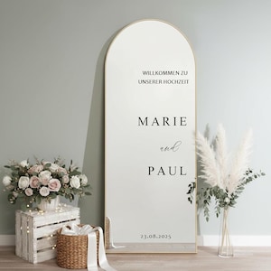 Puede incluir: Un espejo grande y arqueado con marco dorado. El espejo muestra el texto "WILLKOMMEN ZU UNSERER HOCHZEIT", "MARIE and PAUL", y la fecha "23.08.2025". Decoración de boda.