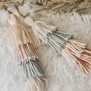 May include: Two multi-colored tassels with a light pink, light blue, and white color scheme. The tassels are made of yarn and are hanging from a white fluffy surface.