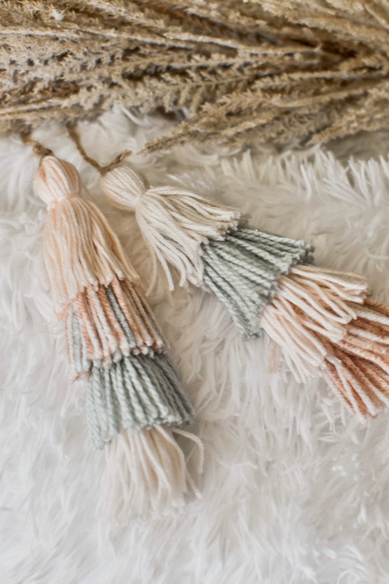 May include: Two multi-colored tassels with a light pink, light blue, and white color scheme. The tassels are made of yarn and are hanging from a white fluffy surface.