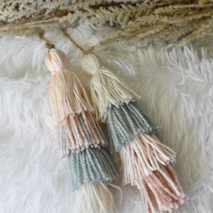 May include: Two multi-colored yarn tassels with a light pink, light blue, and white color scheme. The tassels are hanging from a white fluffy surface.