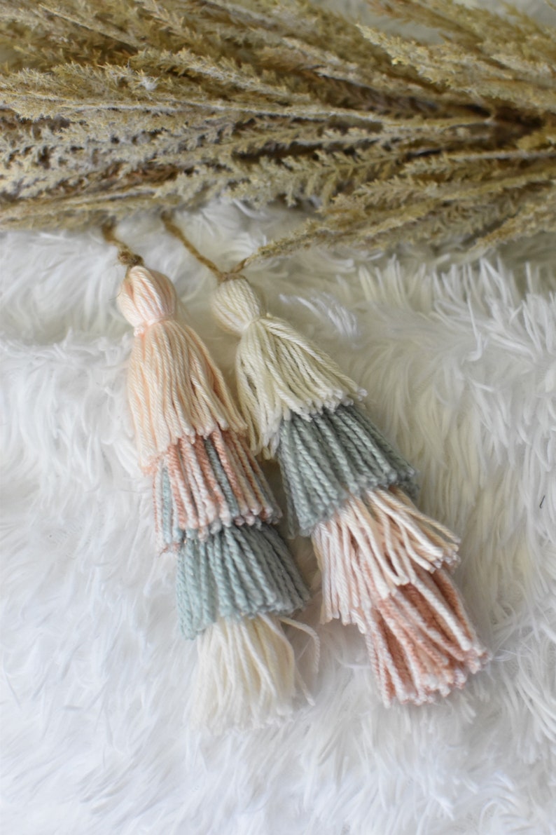 May include: Two multi-colored yarn tassels with a light pink, light blue, and white color scheme. The tassels are hanging from a white fluffy surface.