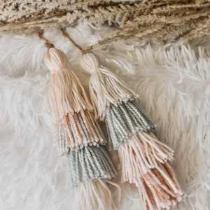 May include: Two multi-colored tassel wall hangings with a light pink, cream, and light green color scheme. The tassels are hanging on a white fluffy surface.
