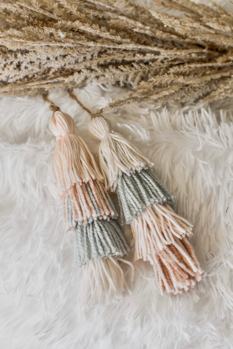May include: Two multi-colored tassel wall hangings with a light pink, cream, and light green color scheme. The tassels are hanging on a white fluffy surface.