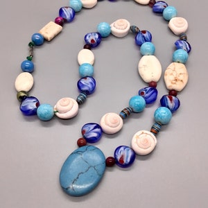 Peut inclure: Un collier artisanal avec un grand pendentif turquoise et diverses perles. Le collier comprend des perles de verre blanches, bleues et rouges, ainsi que des perles en forme de coquillage. Les perles sont enfilées avec une chaîne délicate, créant un accessoire unique et accrocheur.