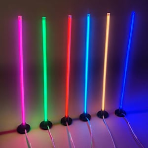 May include: Six vertical LED light sticks standing on circular black bases. Each stick emits a distinct vibrant colour: magenta, green, red, cyan, orange, and blue. Thin power cords extend from the base of each illuminated cylindrical light fixture.