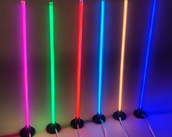 May include: Six vertical LED light sticks standing on circular black bases. Each stick emits a distinct vibrant color: magenta, green, red, cyan, orange, and blue. Thin power cords extend from the base of each illuminated cylindrical light fixture.