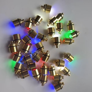 May include: A collection of small, cylindrical LED lights with a metallic gold finish. Each light emits a different color, including red, green, blue, and white. The lights are scattered across a white surface.