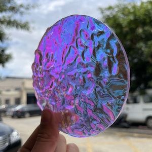 Könnte beinhalten: Ein rundes, irisierendes Glasobjekt mit strukturierter Oberfläche. Das Glas reflektiert Schattierungen von Lila, Blau und Rosa. Das Objekt wird vor einem verschwommenen Hintergrund im Freien gehalten.