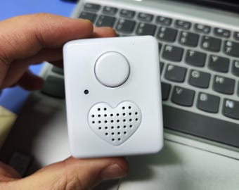 May include: A small, white, square device with a circular button on top and a heart-shaped speaker grill. The device is held in a hand, with a laptop keyboard in the background.