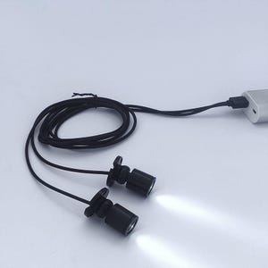 Puede incluir: Dos pequeñas luces LED negras con haces blancos brillantes están conectadas por un cable negro a un cargador USB. Las luces son cilíndricas con una base acampanada. El cable está enrollado.