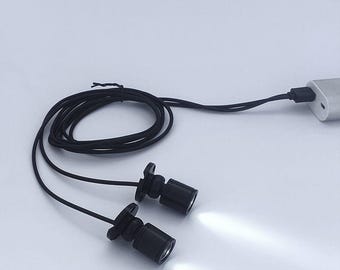 May include: Two small black LED lights with bright white beams are connected by a black cord to a USB charger. The lights are cylindrical with a flared base. The cord is coiled.