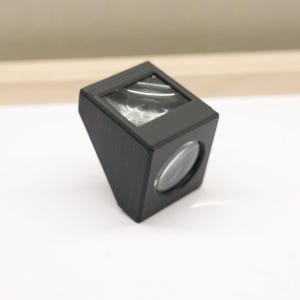 Puede incluir: Un dispositivo rectangular negro, en ángulo, con una lente en la parte delantera y una abertura cuadrada en la parte superior. El dispositivo parece estar hecho de un material negro mate y se encuentra sobre una superficie blanca. La lente es clara y redonda.