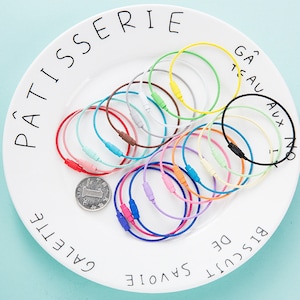 May include: A collection of 18 colorful metal key rings with a silver cable chain. The key rings are arranged in a circle on a white plate with French words printed on the edge.